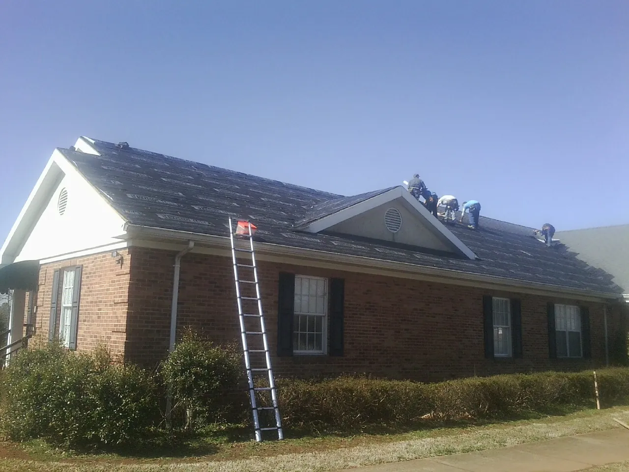 Crew tearing shingles off a brick ranch roof with a ladder set — Emergency Roof Repair work by our crew in Bryceville