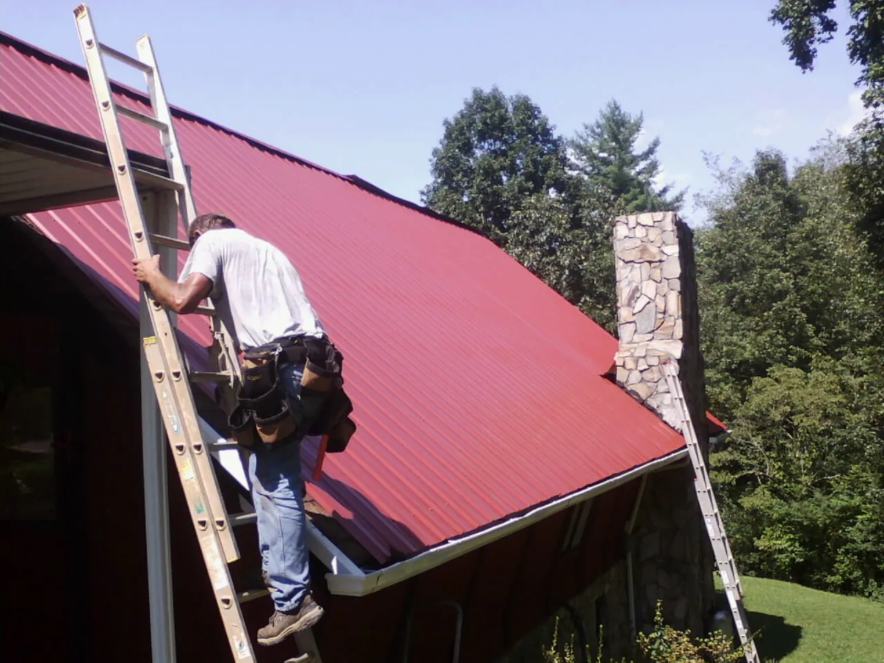 Roofer on a ladder beside a red metal roof and stone chimney — Soffit & Fascia Repair by Roofing Brothers in Bryceville
